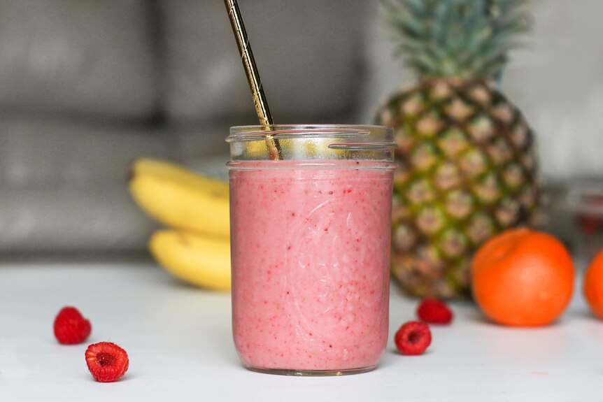 A pink berry smoothie in a mason jar with a metal straw, pictured on a kitchen bench with fresh fruit behind.