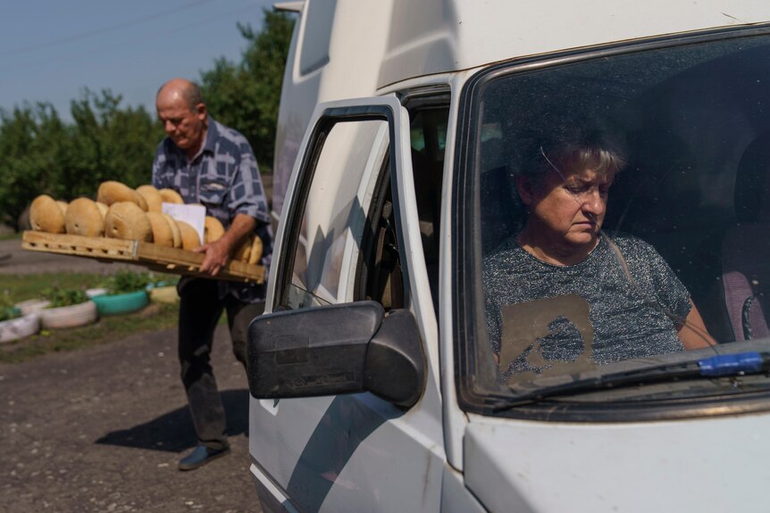 A man carrying a tray of bread behind a van with a woman in the front passenger seat