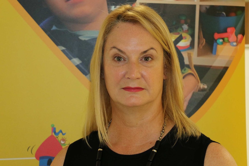 A head and shoulders shot of a middle-aged woman with blonde hair standing in front of a yellow wall.