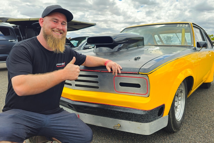 A bearded man kneeling next to a yellow car, he is smiling and giving a thumbs up