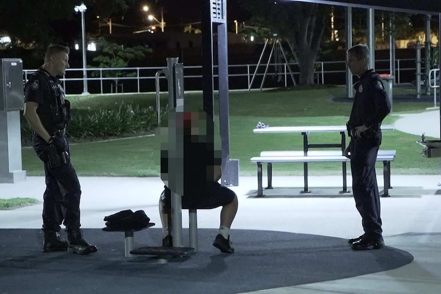 Two police officers stand around a teenage boy who is sitting down in a park at night.