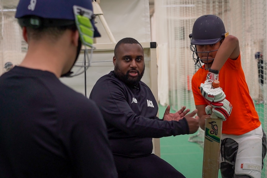 A man teaches two young boys cricket batting techniques.