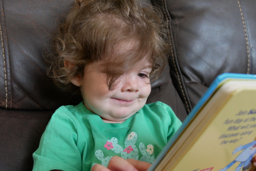 Toddler aged girl sitting on couch reading a book with a smile.
