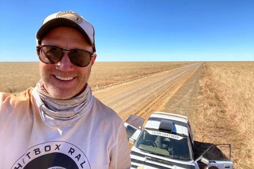 A smiling man in a cap and sunglasses stands on a desert road.