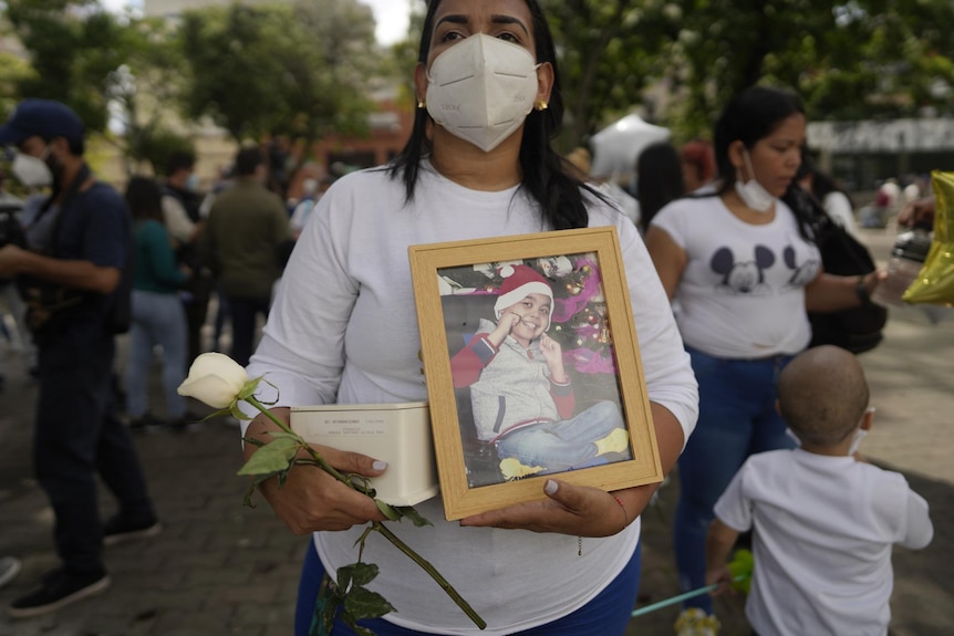 Woman holding a frame photo of her son, an urn with his ashes and a white rose 
