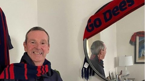 A smiling man ins Melbourne AFL gear stands next to a mirror with a "Go Dees" sign on it. 