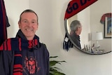 A smiling man ins Melbourne AFL gear stands next to a mirror with a "Go Dees" sign on it. 