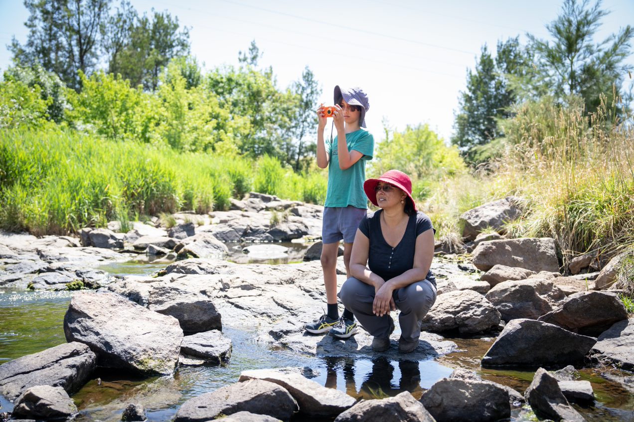 A young boy with a camera and a woman in a pink hat stand on wet river rocks.