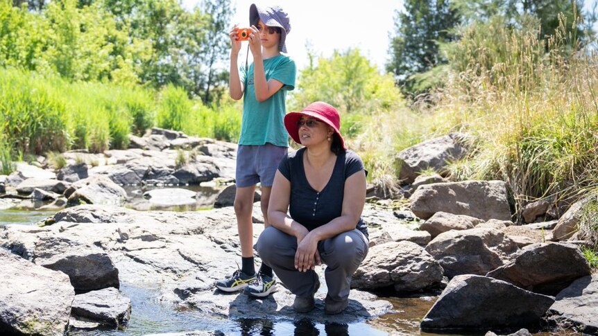 A young boy with a camera and a woman in a pink hat stand on wet river rocks.