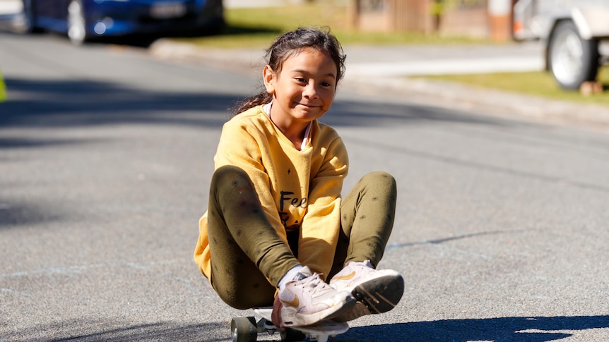 An image of a girl, smiling as she sits on a skateboard rolling down a street in Woodridge