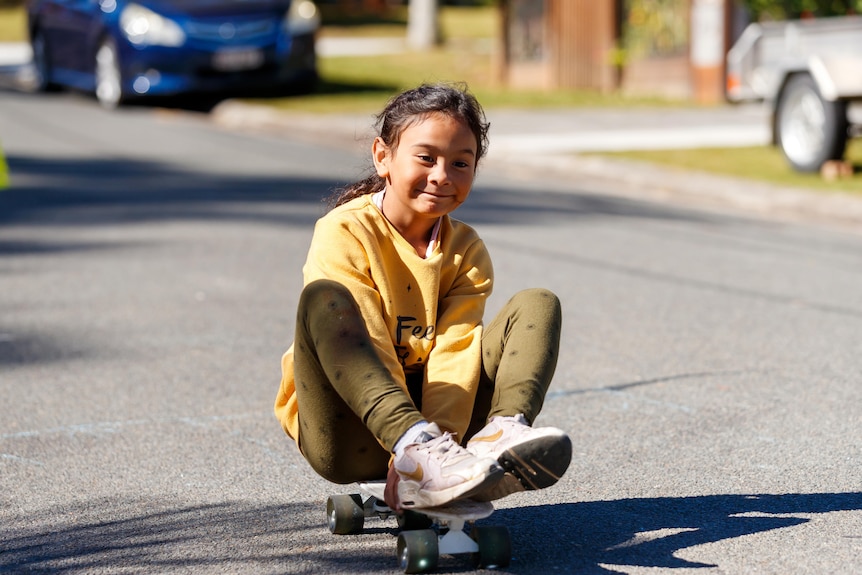 An image of a girl, smiling as she sits on a skateboard rolling down a street in Woodridge