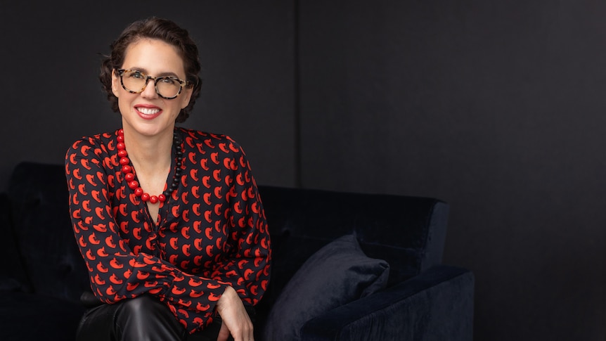 Portrait of a middle-aged woman against a black backdrop. She's wearing a red spotty top and glasses.
