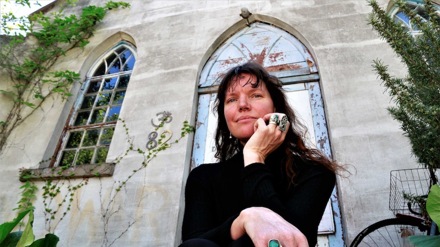 Woman sits outside church, wearing black turtle neck and showing off handmade jewellery.