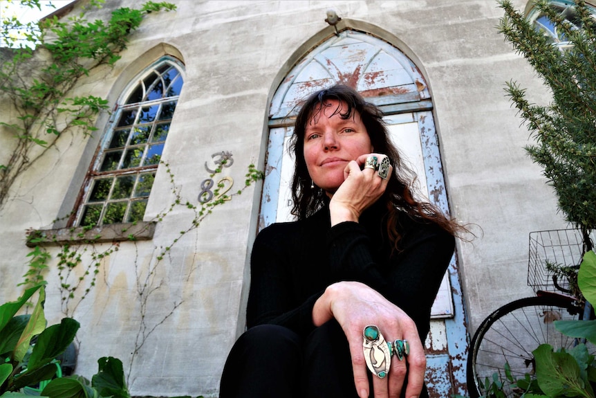 Woman sits outside church, wearing black turtle neck and showing off handmade jewellery.