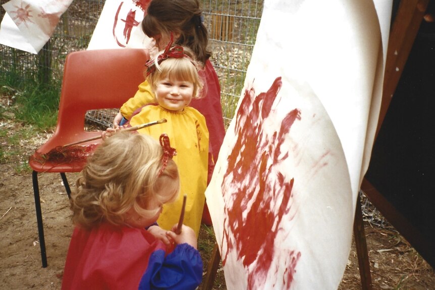 A young girl with blonde hair wears a yellow smock while painting outdoors