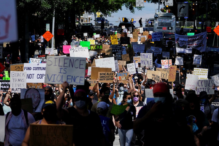 A big crowd of people holding signs walk down a city street, some wearing masks, on sunny afternoon.