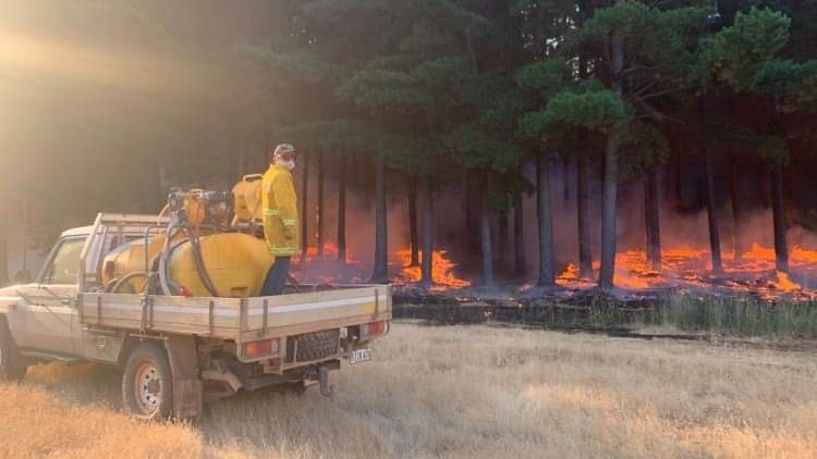 A man stands on the back of a ute dressed in firefighting gear hosing down a fire