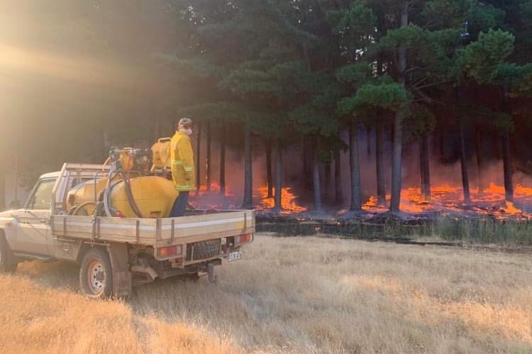 A man stands on the back of a ute dressed in firefighting gear hosing down a fire