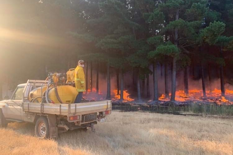 A man stands on the back of a ute dressed in firefighting gear hosing down a fire