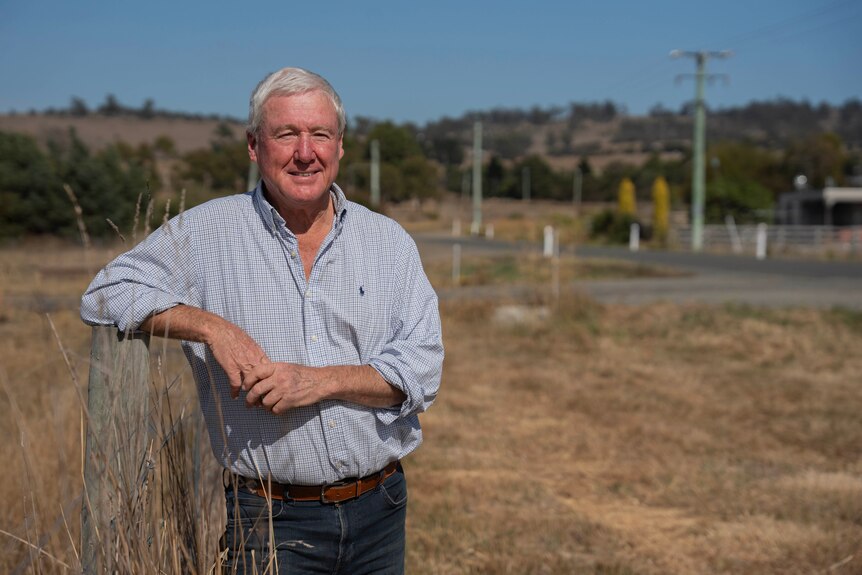 A male politician in a blue shirt stands along the side of a fence in a field.
