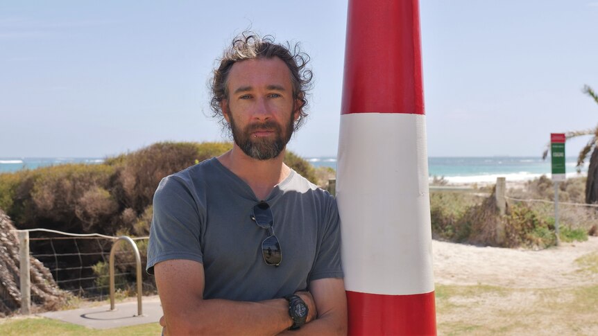 Man leaning on lighthouse pole, folding his arms, with sunny beach in the background