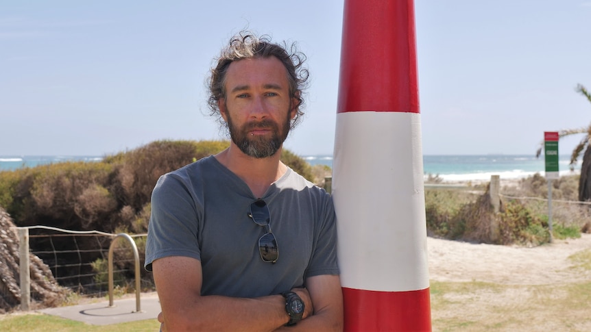 Man leaning on lighthouse pole, folding his arms, with sunny beach in the background