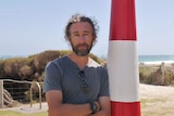 Man leaning on lighthouse pole, folding his arms, with sunny beach in the background