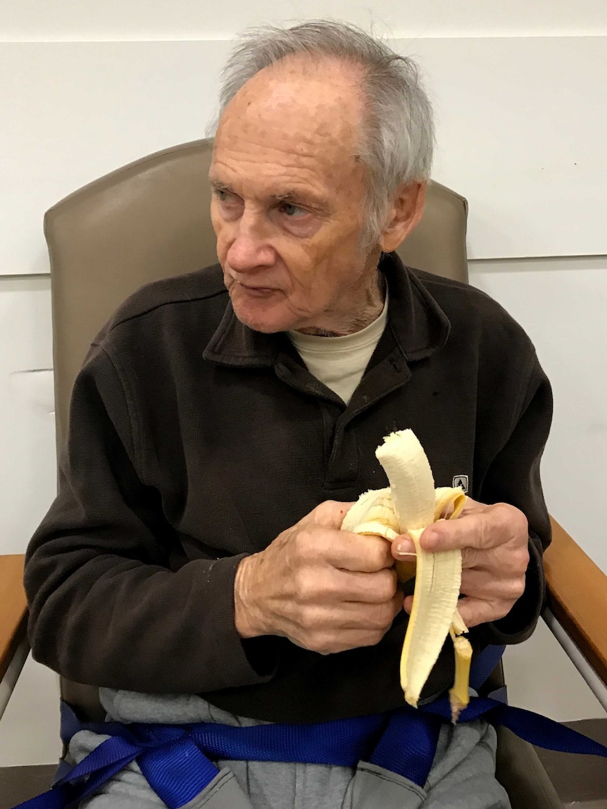 Terry Reeves sits in a chair tied down with a seatbelt-like strap, eating a banana.