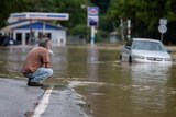 Man sits on side of flood waters as car floats by