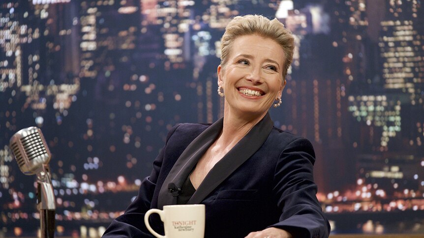 Emma Thompson smiles broadly while seated at desk with mug and vintage microphone in front of nightscape backdrop.