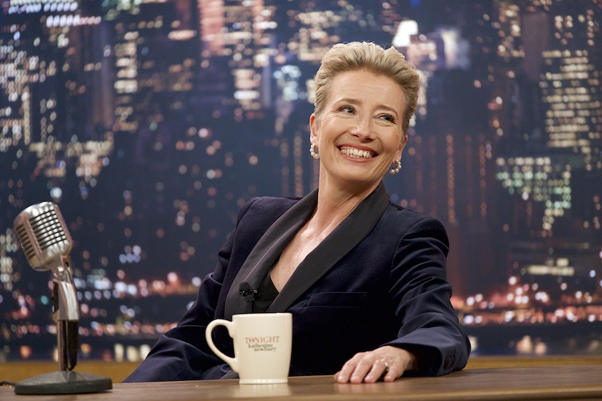 Emma Thompson smiles broadly while seated at desk with mug and vintage microphone in front of nightscape backdrop.