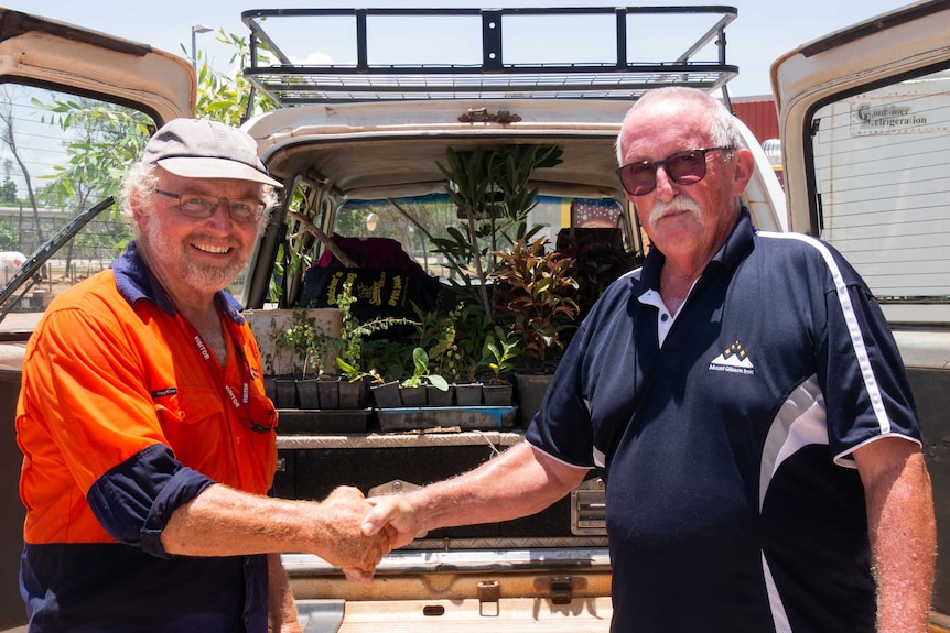 Two men shake hands in front of a car loaded with plants.