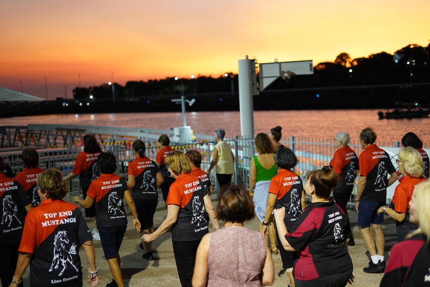 A group of people line dancing in an outdoor area alongside a river, at sunset. 