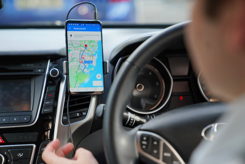 A phone with Google Maps open is mounted on the air vent next to the steering wheel inside a car.