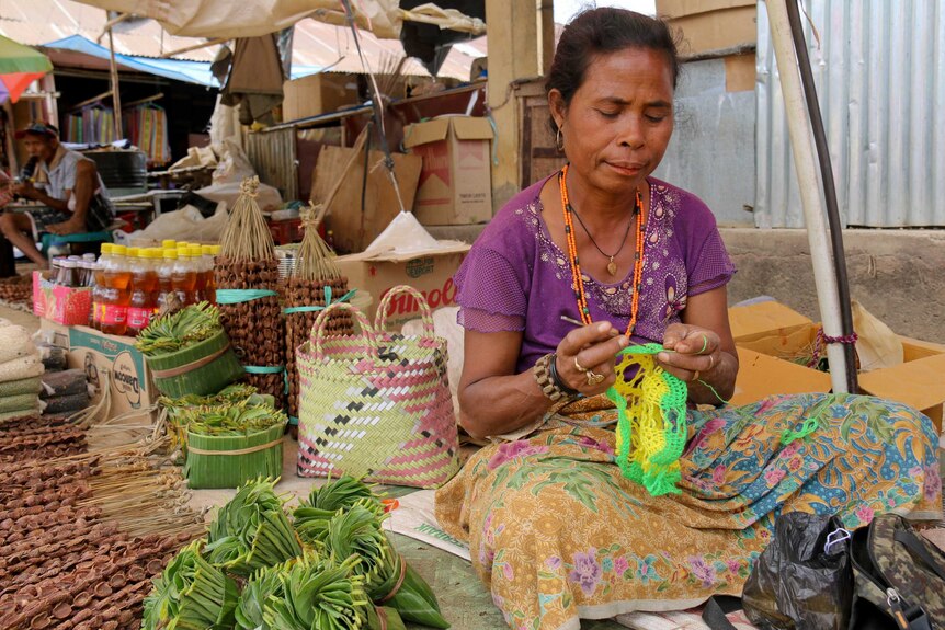 Timorsese woman sitting on a mat at her stall crocheting with green and yellow wool.