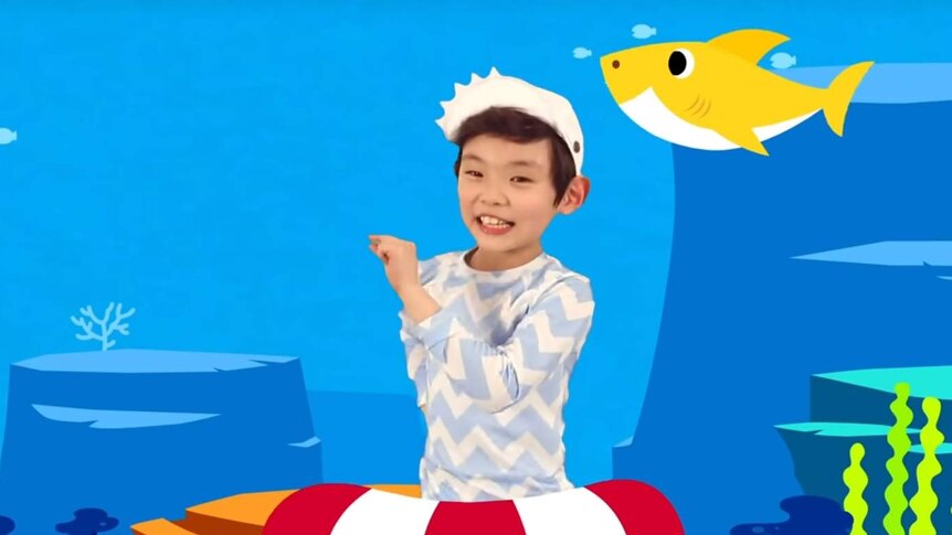 A boy pinches his fingers together in front of a cartoon under-water scene with a cartoon yellow shark over his left shoulder.