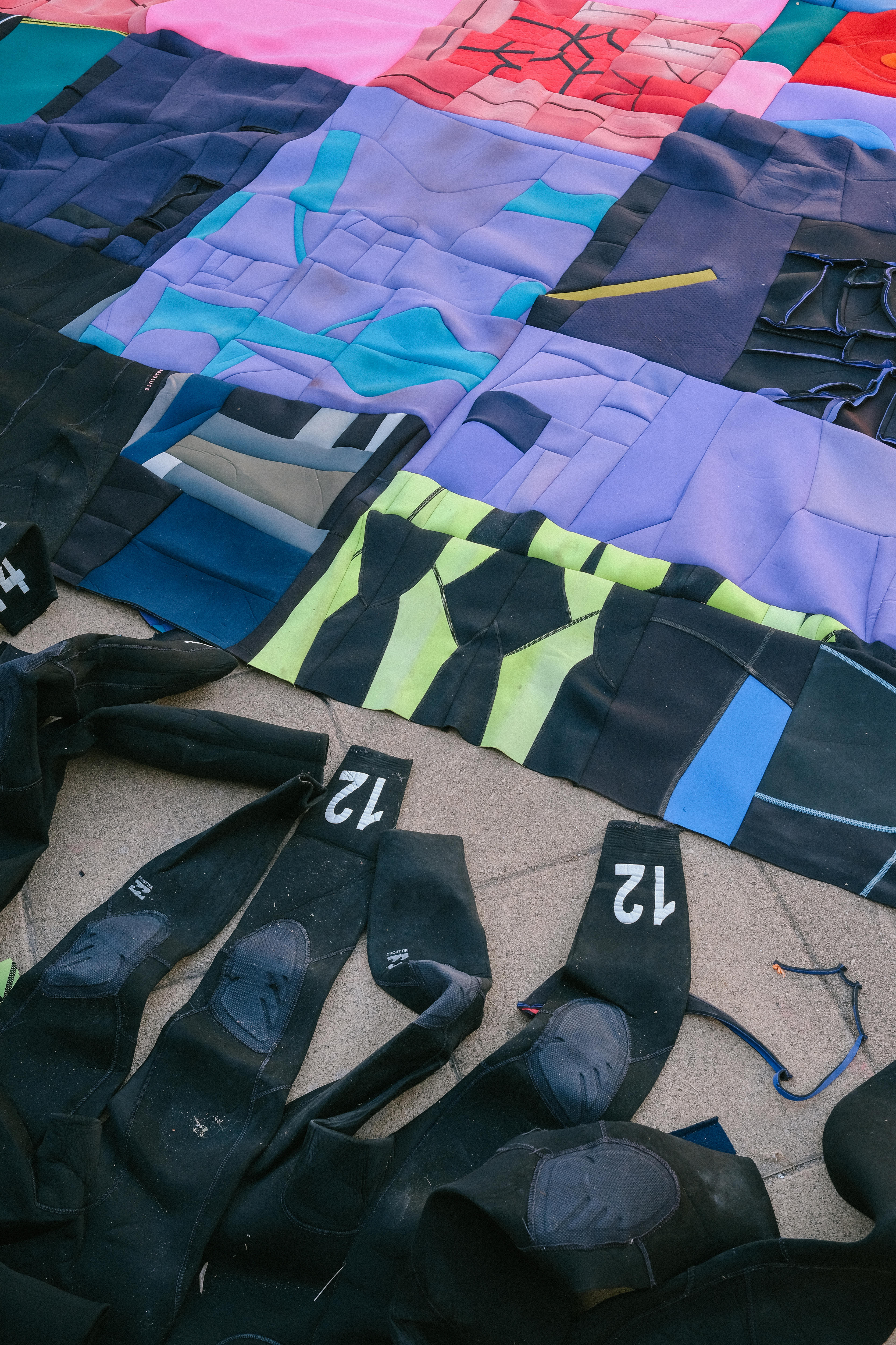 Second hand black wetsuits lay on the ground next to a completed artwork, made out of cut up wetsuits 