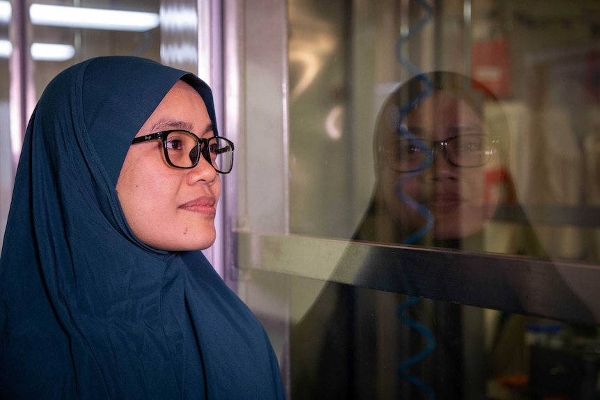 Dr Sri Handayani Irianingsih wears a green hijab and glasses and looks into a lab, her face is reflected on the glass.