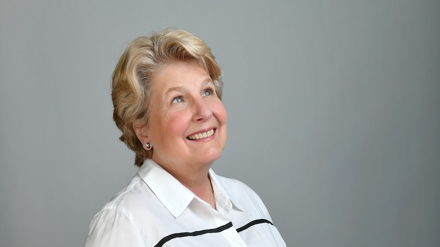 A portrait of Sandi Toksvig looking up and smiling with her hands in her pockets in front of a plain backdrop.