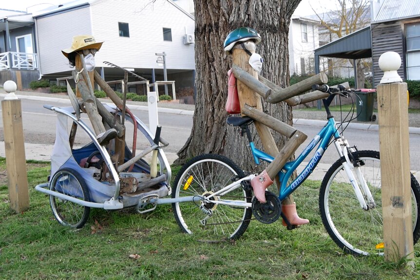 Two treated pine statues sitting on bikes with repair tools.