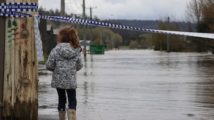 Girl looks at Meander River in flood near pole with police tape.
