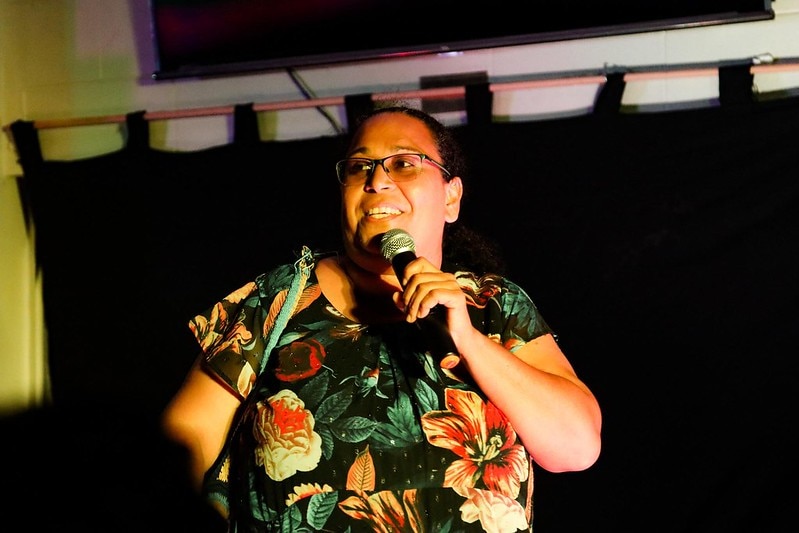 Female Indigenous comedian performing. She's wearing her hair up and brightly coloured dress with hibiscus and other flowers