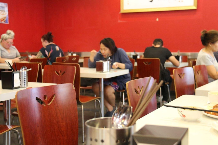 Two people eating and three others sitting at tables on their phones in an otherwise empty restaurant