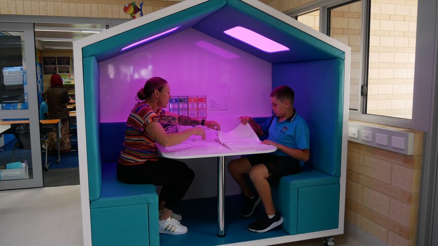 A woman and a boy sit in a large house-shaped pod with purple lighting and workbooks. Behind the pod is a classroom.
