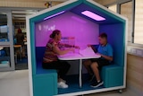 A woman and a boy sit in a large house-shaped pod with purple lighting and workbooks. Behind the pod is a classroom.