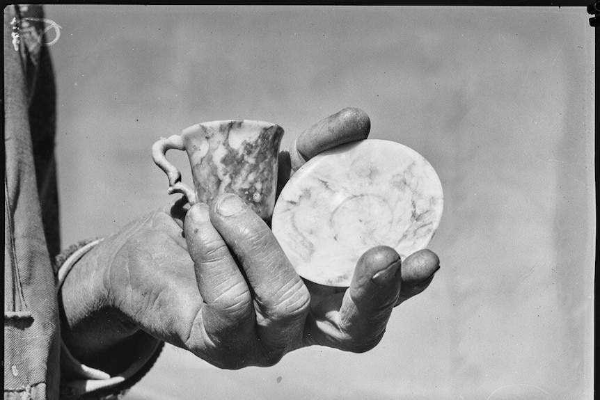 A black and white image of a hand holding a teacup and saucer carved from marble.