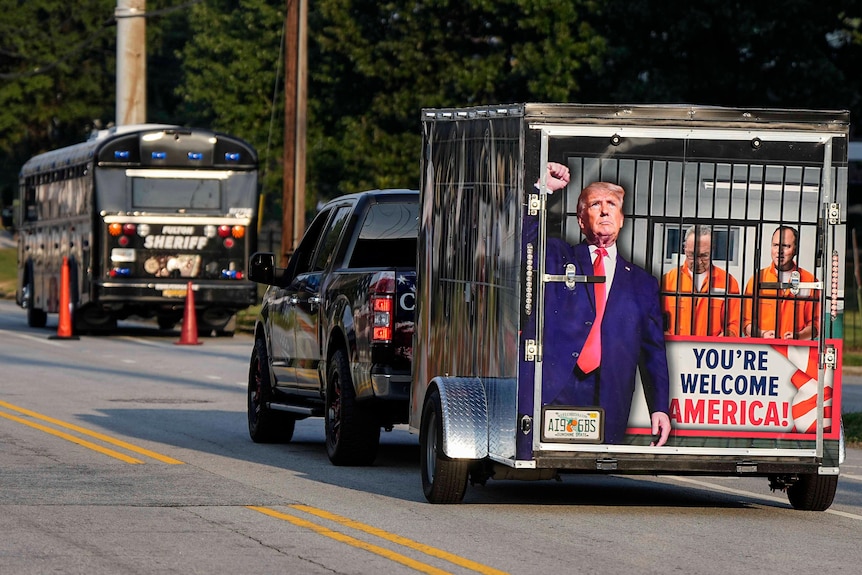 a truck tows a trailer that has an image on the back of it that shows donald trump putting people in prison.