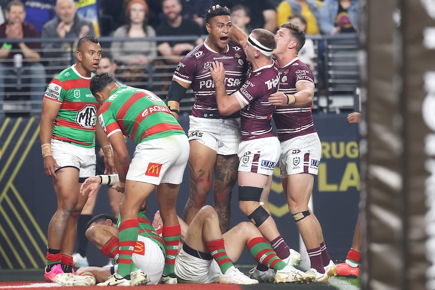 A tall Manly NRL player shouts in celebration as he is patted on the head by teammates while opposing players lie on the ground.