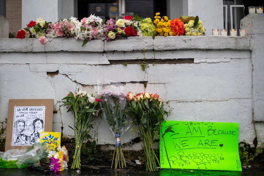 Flowers and signs are displayed at a makeshift memorial in front of a cracked concrete wall.