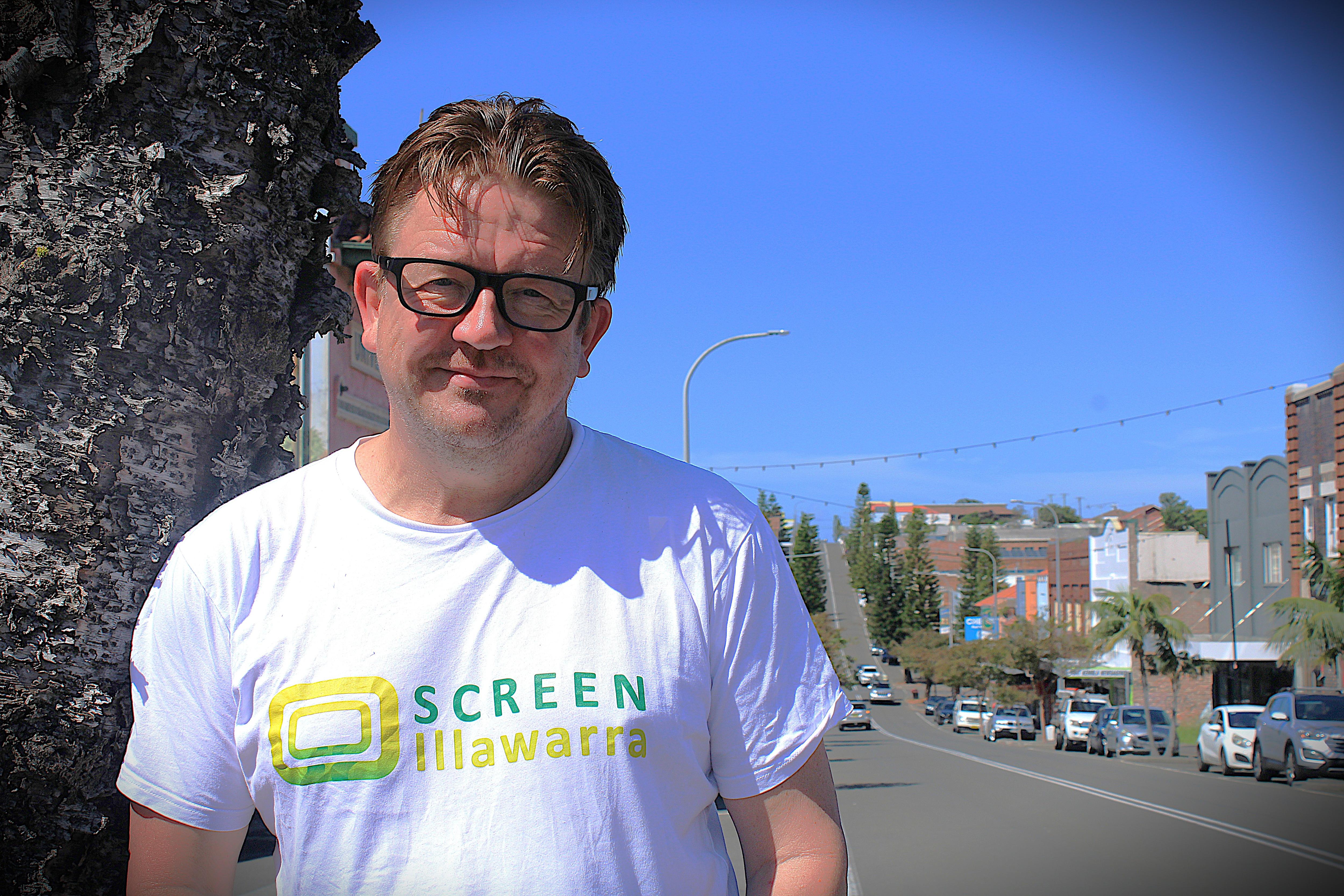 Hombre con cabello moreno y gafas, camisa blanca, sonriendo en la calle principal de Port Kembla
